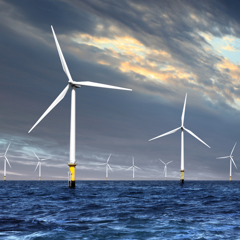 An offshore wind farm with two prominent three-bladed wind turbines standing in choppy blue ocean waters under a dramatic, cloudy sky with hints of sunlight. Additional turbines fade into the hazy background horizon.